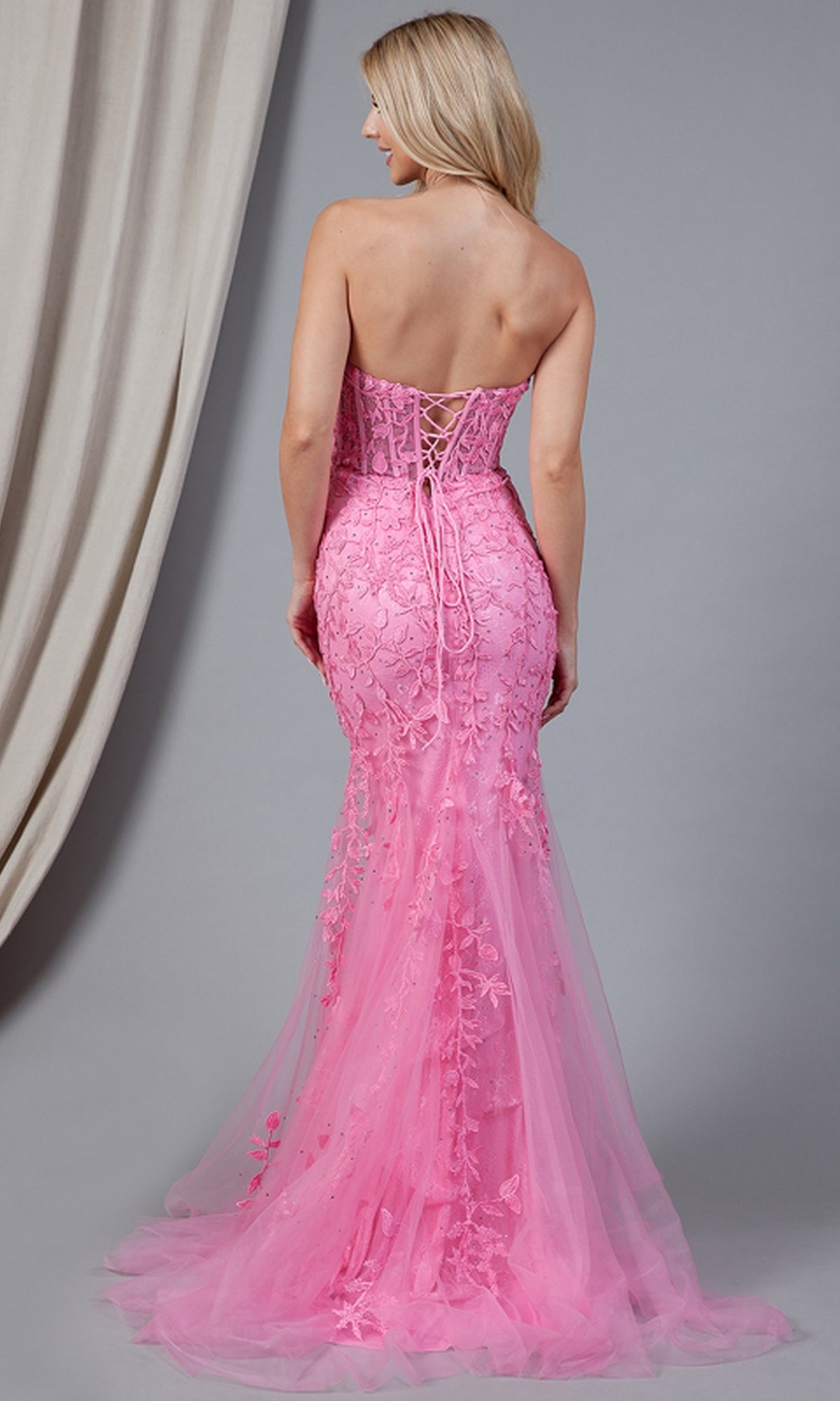 A woman faces away, wearing the Strapless Sweetheart Long Mermaid Prom Dress 7024 with a corset-style bodice and embroidered floral designs, standing before a gray backdrop with a draped curtain.